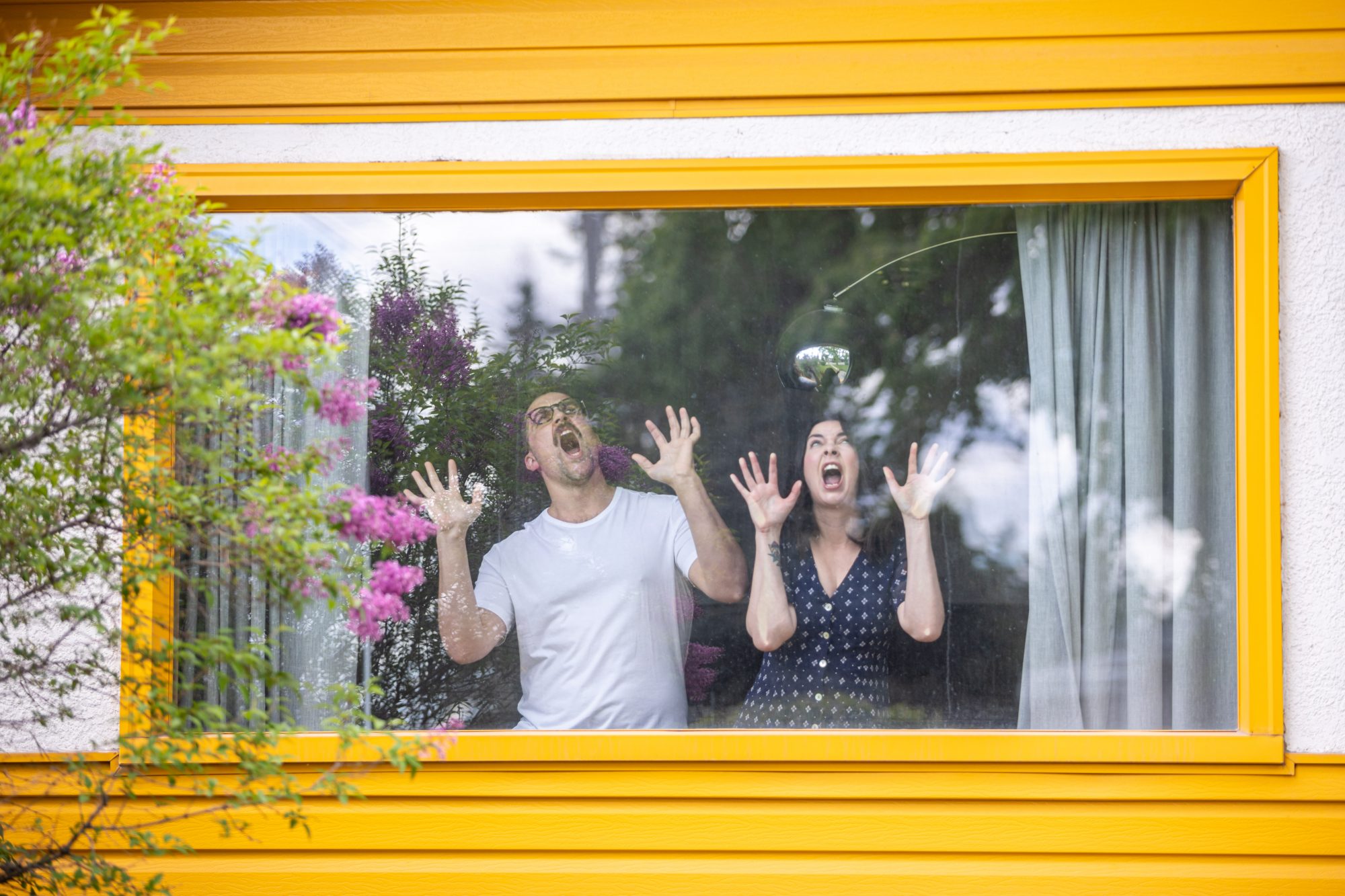 A portrait of a man and a woman making faces while looking out the window of their house