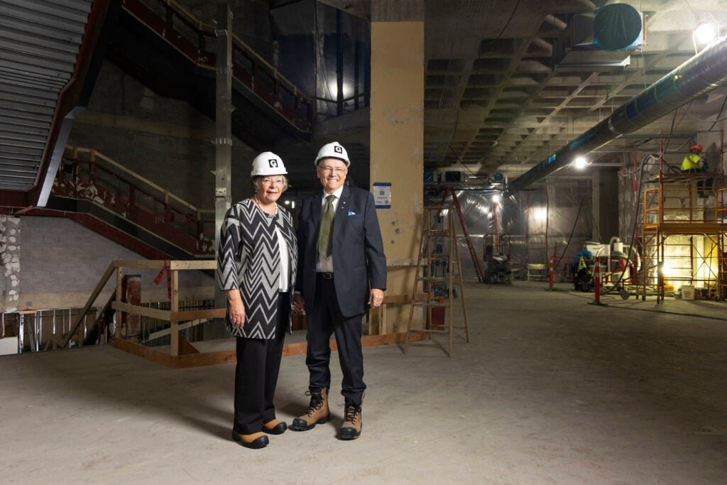 Susan and Dave Werklund stand together on the second floor of Glenbow. Construction continues to their right, while they both wear hard hats in the space.