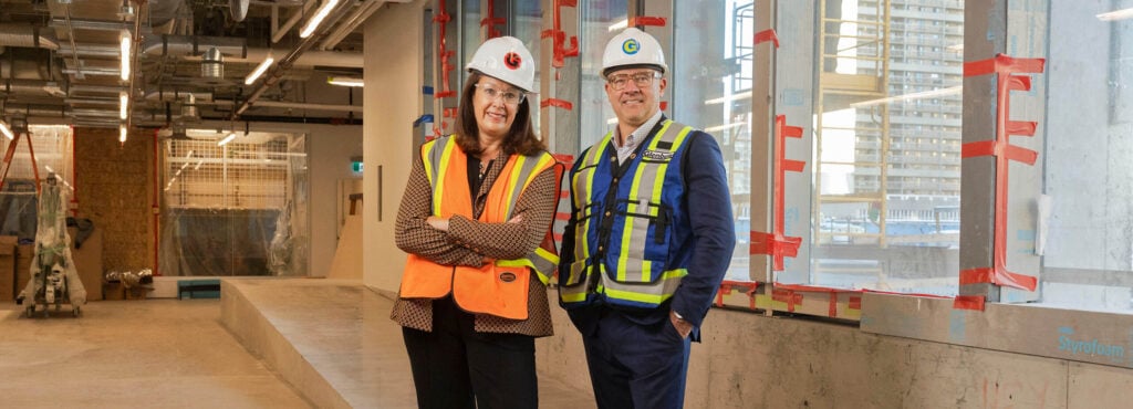 Two people in hard hats and safety vests stand together in a construction zone
