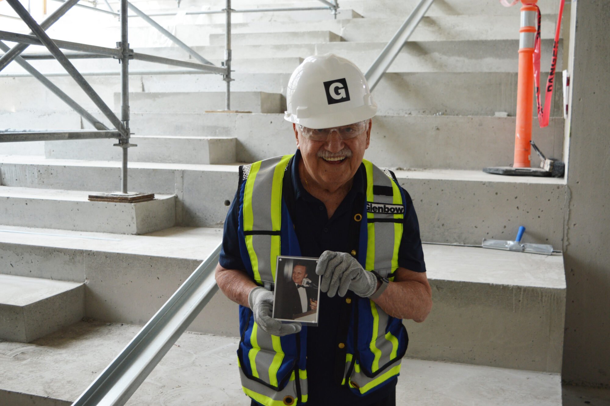 An older man stands in a hardhat, bright safety vest, holding a photo