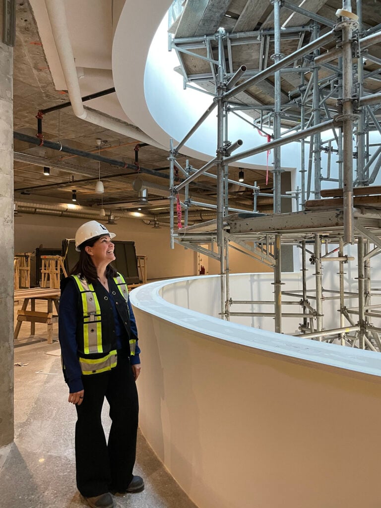 A woman in a hardhat and reflective vest looks up into a skylight partially blocked by scaffolding
