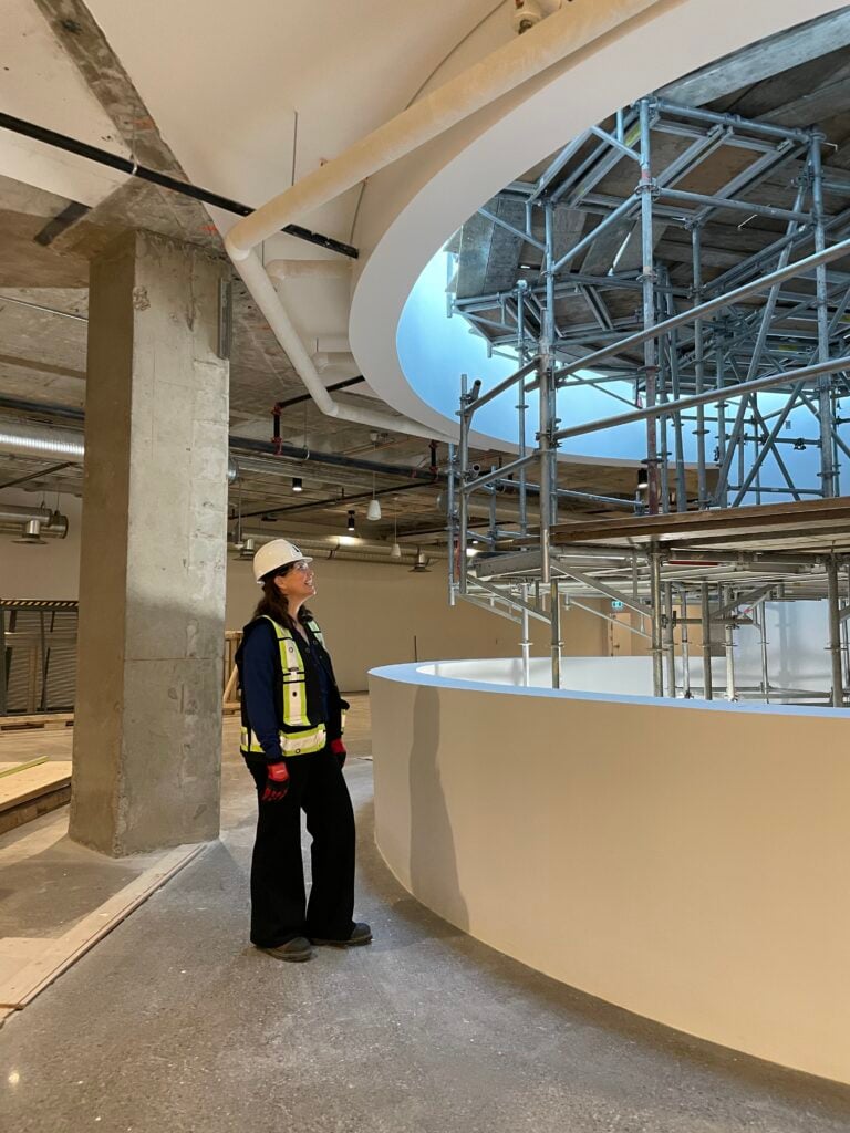 A woman in a safety vest and hard hat looks toward a museum gallery under construction.